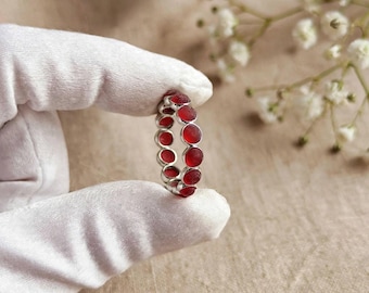 May include: A silver ring featuring a continuous band of small, circular, deep red stones. The ring is held between two fingers covered in white fabric gloves, showcasing the vibrant red gems set within a polished silver metal frame.