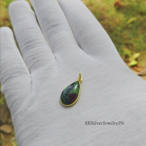 Può includere: Un ciondolo a forma di goccia con una gemma di rubino zoisite lucida con motivi maculati verde scuro e rosso magenta, incastonato in una sottile montatura in metallo dorato con un piccolo occhiello. Il testo SRSilverJewelryIN è visibile.
