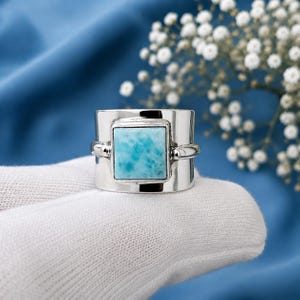 May include: A wide silver band ring featuring a square, polished light blue Larimar gemstone set in the center. The stone displays a marbled, sky-blue pattern with white veining, held securely by a smooth, polished silver bezel and frame.