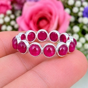 May include: A silver eternity band featuring a continuous row of round, faceted, deep pinkish-red gemstones set in individual polished silver bezels. The ring displays a uniform circular design with each stone securely held in a metallic frame.