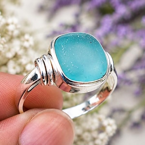 May include: A silver ring featuring a smooth, square-shaped teal sea glass stone set in a polished metal bezel. The band is accented with three horizontal wire wraps on each side of the setting, creating a textured detail along the shank.