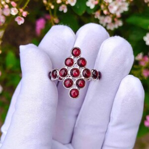 Könnte beinhalten: Ein Silberring mit einer Anordnung von zehn runden, tiefroten Granat-Cabochon-Steinen in Rautenform. Die polierte Metallfassung hält jeden Stein sicher in einer Zargenfassung und erzeugt so ein symmetrisches und geometrisches Design.