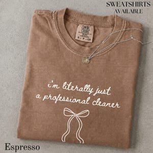 以下が含まれることがあります： クルーネックとComfort Colorsのラベルが付いた茶色の半袖Tシャツ。シャツには「i'm literally just a professional cleaner」という白い文字と、リボンのシンプルな白い線画が描かれています。生地の上にはシルバーのチェーンネックレスが置かれています。