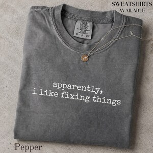 May include: A charcoal gray short-sleeve t-shirt with a crew neckline and the white text "apparently, i like fixing things" printed on the front. The shirt features a Comfort Colors brand label inside the collar and a thin gold-tone necklace.