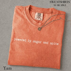 以下が含まれることがあります： クルーネックのヤム色のTシャツで、胸元に「powered by sugar and spite」という白い文字がプリントされています。シャツの内側の襟元にはComfort Colorsのブランドラベルがあり、細い銀色のチェーンネックレスが添えられています。