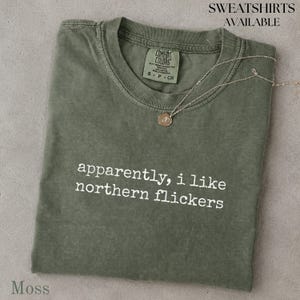 May include: A moss green crew neck t-shirt featuring the white text "apparently, i like northern flickers" printed on the chest. A thin gold-toned chain necklace rests on the neckline, and a brand label is visible inside the collar.
