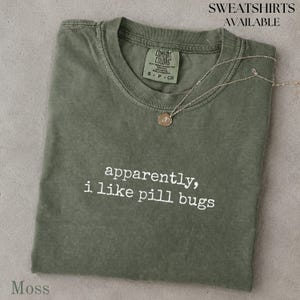 May include: A moss green short-sleeve t-shirt with a crew neckline and white printed text that reads, apparently, i like pill bugs. The shirt features a size small tag inside the collar and a thin gold-toned chain necklace resting on top.