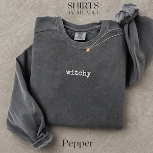 Könnte beinhalten: Ein anthrazitfarbenes Sweatshirt mit Rundhalsausschnitt und Vintage-Waschung. Das Wort "witchy" ist in weißem Garn mittig auf der Brust eingestickt. Eine dünne goldfarbene Kette mit einem kleinen runden Anhänger liegt am Halsausschnitt.