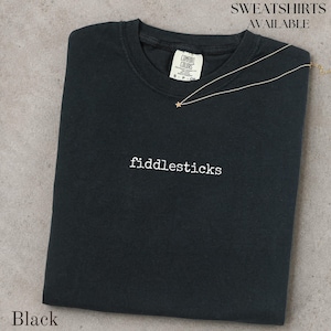 May include: A black short-sleeve t-shirt featuring the word fiddlesticks embroidered in white lowercase letters across the center chest. The shirt has a crew neckline with a Comfort Colors brand label and a thin gold chain necklace with a star charm.