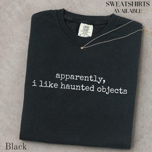May include: A black t-shirt featuring the white text "apparently, i like haunted objects" printed on the front. The shirt has a crew neckline and a white interior neck label. A thin gold-tone necklace with a small star charm rests on the fabric.
