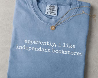 May include: A blue jean colored t-shirt with a crew neckline and the white text "apparently, i like independant bookstores" printed on the front. A gold chain necklace rests on the collar. The inner label reads "Comfort Colors" and "S P CH".