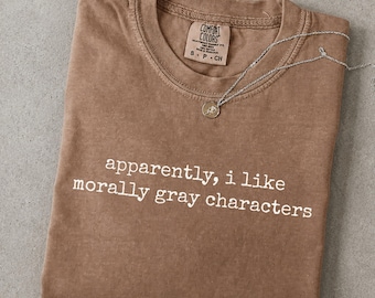 May include: A brown short-sleeve t-shirt with the white text "apparently, i like morally gray characters" printed on the front. The shirt features a Comfort Colors brand label at the inner neckline and a thin silver-toned chain necklace.