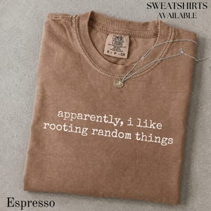 May include: A brown short-sleeved t-shirt with a crew neckline and the white text "apparently, i like rooting random things" printed on the front. A silver-toned necklace with a small circular pendant rests on the collar. The shirt has a tag.