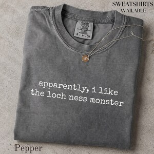 May include: A charcoal gray crew neck t-shirt featuring the white text "apparently, i like the loch ness monster" printed on the front. The shirt has a Comfort Colors brand label inside the collar and is displayed with a thin gold chain necklace.