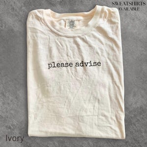 May include: An ivory-coloured short-sleeve t-shirt featuring the text "please advise" printed in black across the centre of the chest. The shirt has a crew neckline and a Comfort Colors brand label visible on the inside of the collar area.