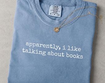May include: A blue jean colored t-shirt with a crew neckline and a white graphic print that reads, "apparently, i like talking about books." A gold-toned chain necklace with a small circular pendant rests on the fabric near the collar.