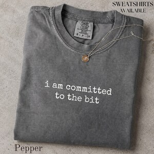 May include: A charcoal grey crew neck t-shirt with a vintage-style wash, featuring the white printed text "i am committed to the bit" across the chest. A delicate gold-toned chain necklace with a small circular pendant rests on the neckline.