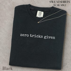 May include: A black crewneck t-shirt featuring the white text "zero tricks given" printed across the chest. The shirt has a Comfort Colors brand label at the inner neckline. A thin gold-toned necklace with a small star charm rests on the fabric.