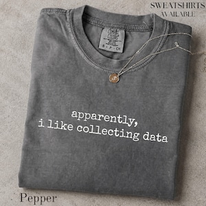 May include: A charcoal gray crew neck t-shirt with a washed-out finish. The text "apparently, i like collecting data" is printed in white across the chest. A thin gold-toned necklace with a small circular pendant rests on the neckline of the shirt.