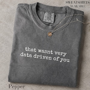May include: A charcoal grey short-sleeve t-shirt with a crew neckline and the white text that reads, that wasnt very data driven of you. A thin gold-tone chain necklace with a small circular pendant rests on the collar. The shirt features a Comfort Colors brand label inside the neck.