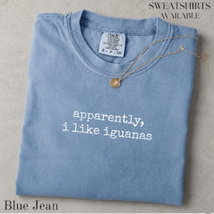 Puede incluir: Una camiseta de color azul vaquero con el texto blanco "apparently, i like iguanas" impreso en la parte delantera. La camiseta cuenta con una etiqueta de la marca Comfort Colors en el cuello interior y se muestra con un collar de cadena fina dorada.