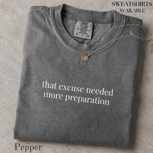 Könnte beinhalten: Ein anthrazitfarbenes T-Shirt mit Rundhalsausschnitt und Vintage-Waschung, das auf der Brust den weißen Schriftzug "that excuse needed more preparation" trägt. Eine zarte goldfarbene Kette mit kleinem runden Anhänger liegt am Ausschnitt.