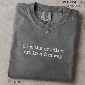 Könnte beinhalten: Ein anthrazitfarbenes Kurzarm-T-Shirt mit Rundhalsausschnitt und dem weißen Text "i am the problem but in a fun way" auf der Brust. Eine dünne goldfarbene Kette mit einem kleinen runden Anhänger liegt auf dem Kragen des T-Shirts.