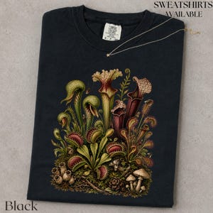 May include: A black short-sleeved t-shirt featuring a detailed graphic print of various carnivorous plants, including pitcher plants and Venus flytraps, in shades of green, red, and brown, with a small white tag at the inner neckline.