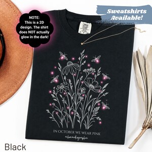 Peut inclure: Un t-shirt noir avec une illustration au trait blanc de fleurs sauvages et de lucioles avec des accents roses lumineux. Le texte indique : NOTE: This is a 2D design. The shirt does NOT actually glow in the dark! IN OCTOBER WE WEAR PINK echocardiographer.