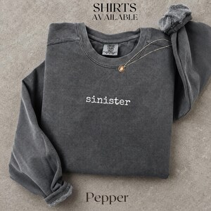May include: A charcoal gray long-sleeve crewneck sweatshirt with the word sinister embroidered in white lowercase letters on the chest. The garment features a vintage-washed finish, a ribbed collar, and a Comfort Colors brand label inside the neck.
