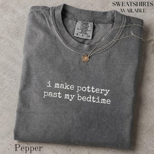 May include: A charcoal gray short-sleeve t-shirt with a crew neckline and a Comfort Colors brand label. The shirt features the white printed text 'i make pottery past my bedtime' across the chest and is accompanied by a thin gold-tone chain necklace.