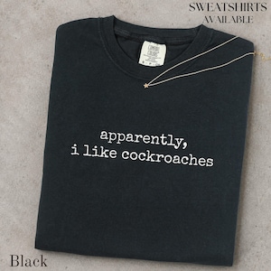 May include: A black crew neck t-shirt featuring the white embroidered text "apparently, i like cockroaches" on the chest. The shirt has a Comfort Colors brand label at the inner neckline and is accompanied by a thin gold chain with a small star charm.
