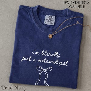 Könnte beinhalten: Ein marineblaues Kurzarm-T-Shirt mit einem weißen Grafikdruck, der "i'm literally just a meteorologist" über einer einfachen Strichzeichnung einer Schleife zeigt. Eine goldfarbene Halskette mit einem kleinen runden Anhänger liegt auf dem Kragen.
