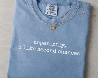 May include: A blue jean colored short-sleeve t-shirt with a crew neckline. The shirt features the white text "apparently, i like second chances" printed on the front. A gold-toned necklace rests on the collar. Text at the top reads "SWEATSHIRTS AVAILABLE".