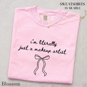 May include: A light pink t-shirt featuring the black text "i'm literally just a makeup artist" above a simple black line drawing of a bow. The shirt has a crew neckline and a Comfort Colors brand label on the inside of the collar.