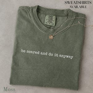 Peut inclure: Un t-shirt à manches courtes vert mousse avec un col rond et le texte blanc be scared and do it anyway imprimé sur le devant. Un fin collier à chaîne dorée avec un petit pendentif circulaire repose sur la zone du col du t-shirt.