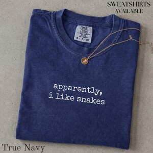 Puede incluir: Una camiseta azul marino de manga corta con cuello redondo y etiqueta de la marca Comfort Colors. La prenda presenta el texto bordado en blanco que dice "apparently, i like snakes". Un collar fino de tono dorado con un pequeño colgante redondo descansa encima.