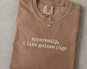 May include: An espresso-colored crew neck t-shirt with a vintage-style wash. The shirt features the white text 'apparently, i like guinea pigs' printed on the chest. A silver-toned chain necklace with a small circular pendant rests on the neckline.