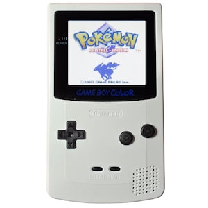 Può includere: Una console portatile Game Boy Color bianca con un pad direzionale nero, due pulsanti azione neri e uno schermo che mostra il logo Pokémon Kristall-Edition con una sagoma blu di Suicune e il testo 2001 GAME FREAK inc.