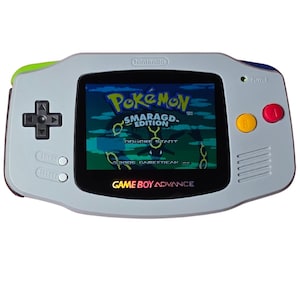 Op de afbeelding: Een lichtgrijze Game Boy Advance handheld console met een zwarte D-pad, gele B-knop en rode A-knop. Het scherm toont het titelscherm van Pokémon Smaragd Edition met de tekst Drücke Start en 2005 Gamefreak Inc.