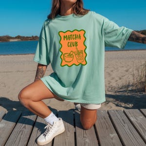 May include: A light mint green t-shirt featuring a rectangular orange graphic with a wavy dark green border. The graphic contains the text "MATCHA CLUB" in dark green letters above an illustration of a matcha bowl with a whisk and an iced drink.