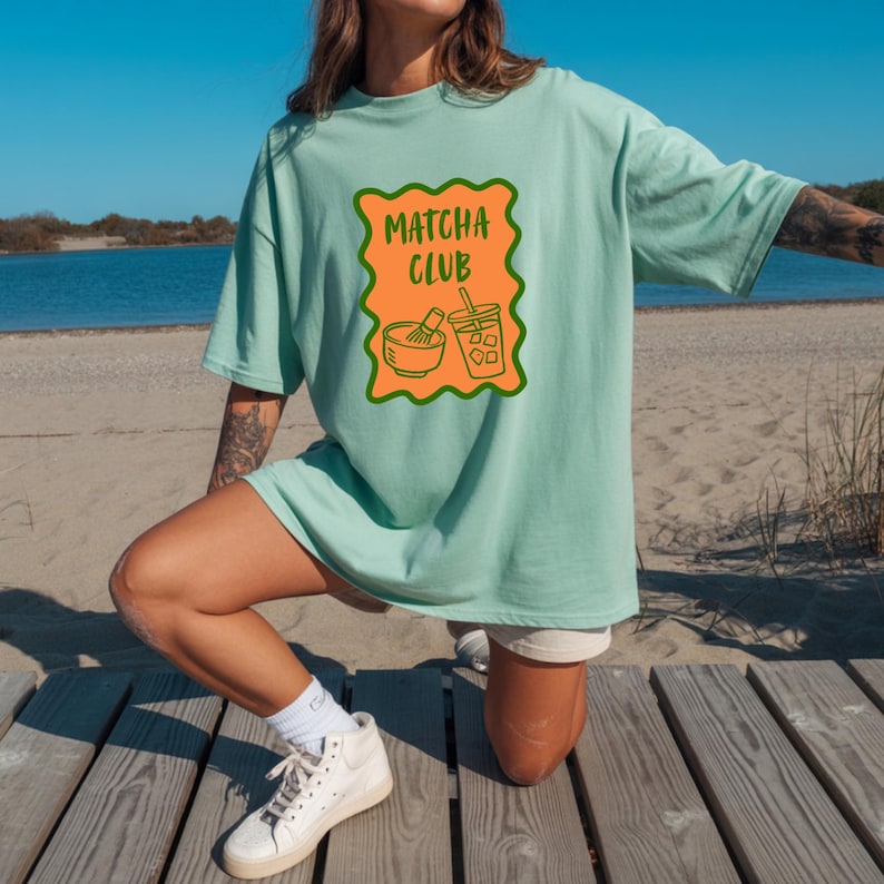 May include: A light mint green t-shirt featuring a rectangular orange graphic with a wavy dark green border. The graphic contains the text "MATCHA CLUB" in dark green letters above an illustration of a matcha bowl with a whisk and an iced drink.