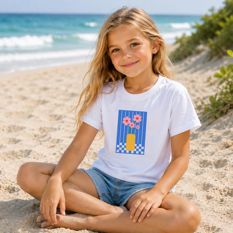 May include: A white short-sleeved t-shirt featuring a graphic print of a yellow vase containing pink flowers against a blue striped background with a blue and white checkered pattern at the base, paired with blue denim shorts.