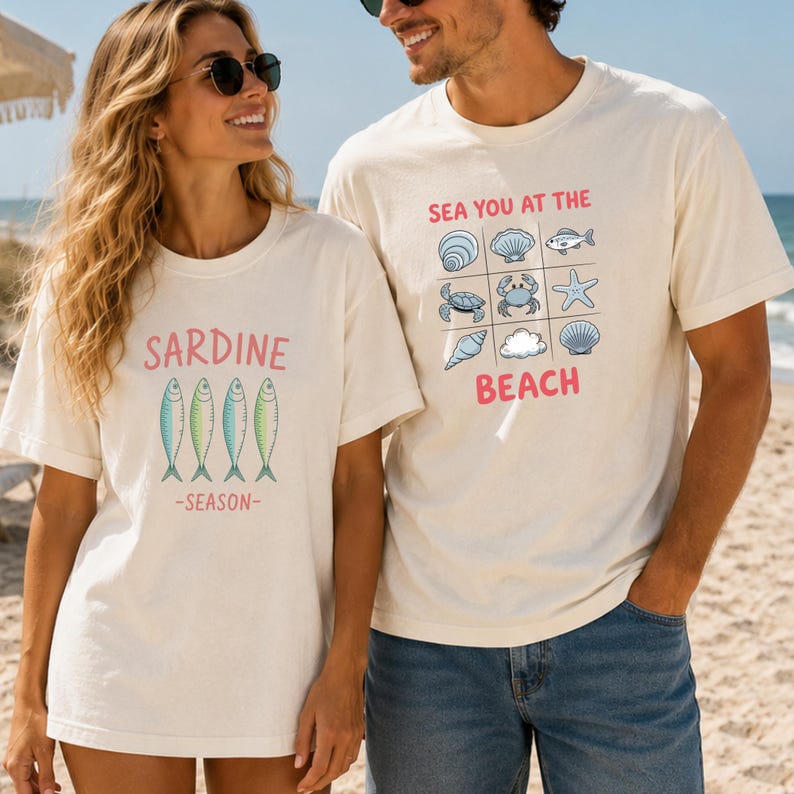 May include: Two off-white short-sleeve t-shirts. One features four green fish illustrations with the pink text SARDINE SEASON. The other displays a grid of nine marine life icons including shells, a fish, a crab, and a starfish with the pink text SEA YOU AT THE BEACH.