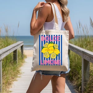 May include: A beige canvas tote bag featuring a graphic print of yellow lemons with green leaves centered over vertical blue stripes. The text DOLCE is printed in pink above the lemons, and LIMONE is printed in pink below the lemons.