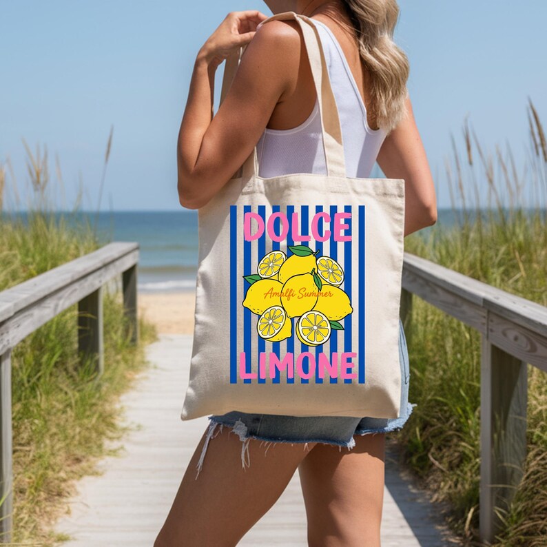 May include: A beige canvas tote bag featuring a graphic print of yellow lemons with green leaves centered over vertical blue stripes. The text DOLCE is printed in pink above the lemons, and LIMONE is printed in pink below the lemons.