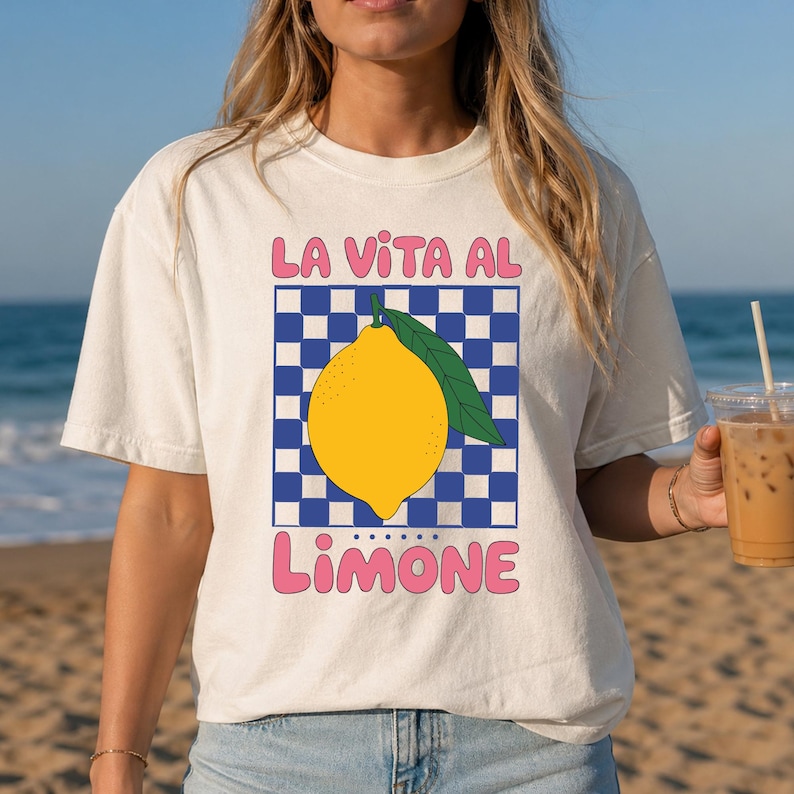 May include: An off-white t-shirt featuring a graphic of a yellow lemon with a green leaf centered over a blue and white checkered square. The pink text above reads LA VITA AL and the pink text below reads LIMONE in a rounded, bold font style.