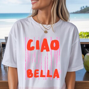 May include: A white short-sleeved t-shirt featuring the text CIAO BELLA in bold orange letters. The text is layered over a background of vertical pink brushstroke lines. The shirt has a crew neckline and a relaxed fit, providing a casual appearance.