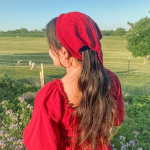 May include: A red knitted head covering with a textured pattern, worn with a red dress. The image is taken outdoors with a green field and blue sky in the background. The head covering is a stylish accessory.