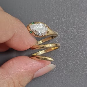 May include: A gold-coloured ring in the shape of a snake. The snake's head features a pear-shaped, clear gemstone and two small green gemstones. The ring coils around the finger in a spiral design.